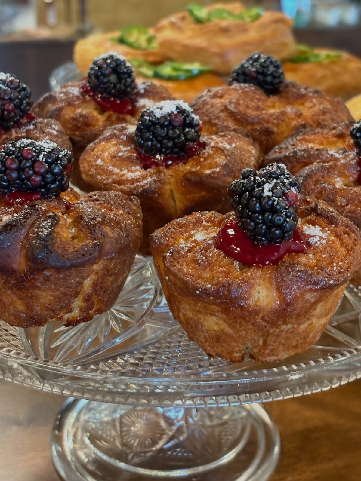 A tray of golden cruffins topped with cream and fresh blackberries on a glass cake stand